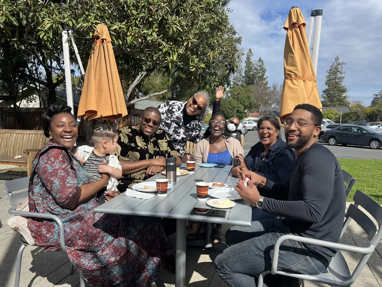 People smile together around an outdoor table, with an adult holding a child.