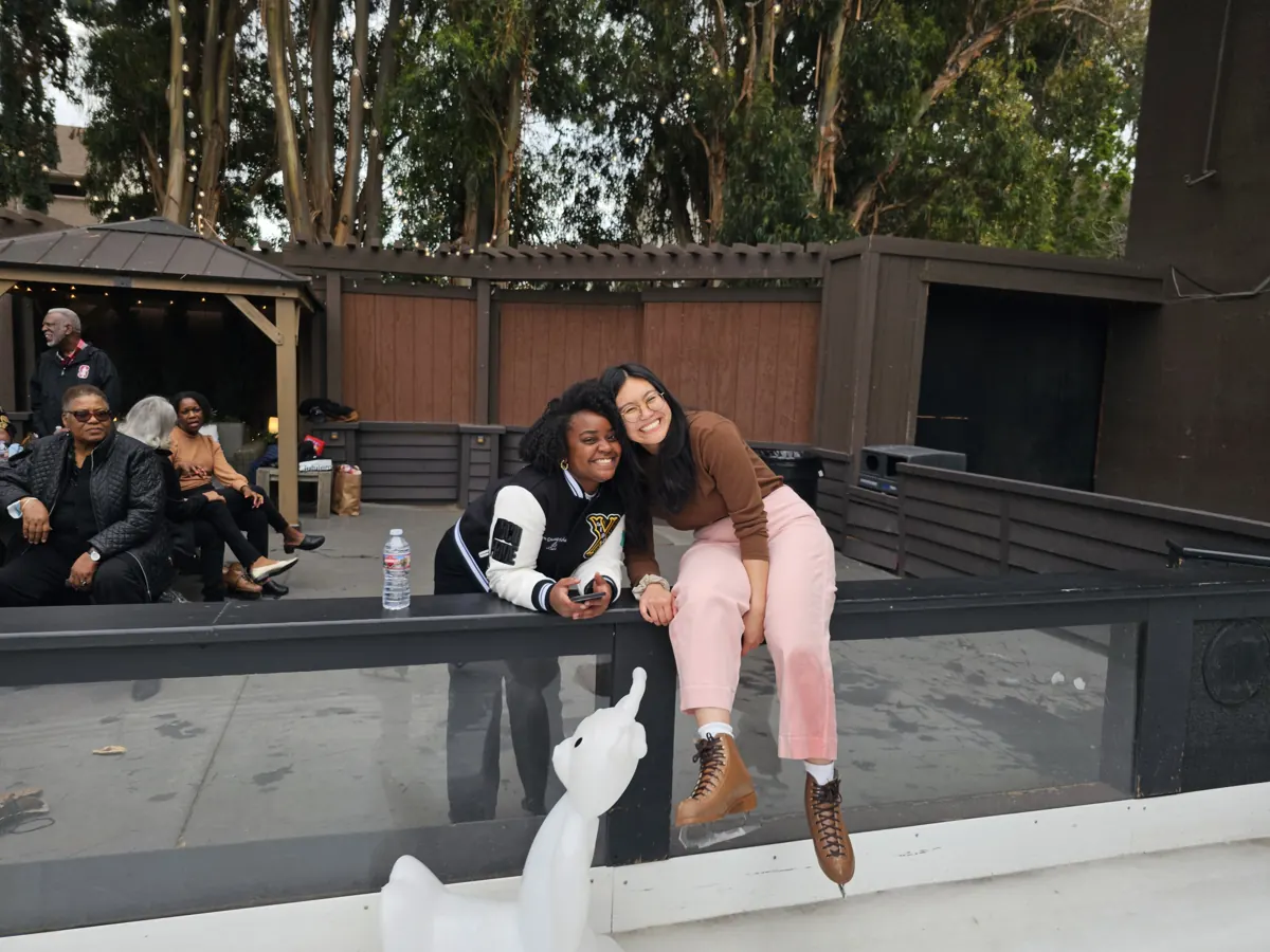 Two friends smile together beside an outdoor ice rink.